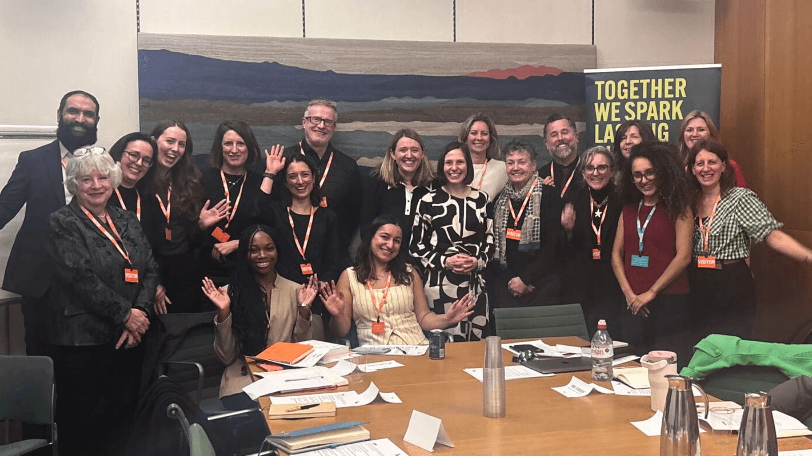 A group of around twenty adults stand and sit closely together around a conference table, smiling and waving at the camera.